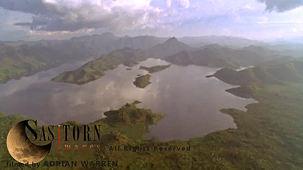 Forward tracking aerial shot, Rwandan countryside, camera passes low over Lake Ruhondo and surrounding hills, cloud formations reflected in the water, camera makes low pass over large ridge covered in patchwork fields