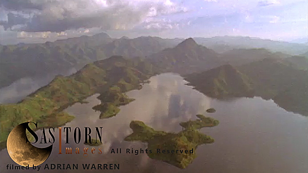 Forward tracking aerial shot, Rwandan countryside, camera passes low over Lake Ruhondo and surrounding hills, cloud formations reflected in the water, camera makes low pass over large ridge covered in patchwork fields