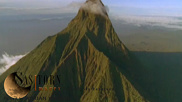 Forward tracking aerial shot, Virunga volcanoes, Mt Mikeno in late afternoon sun with light cloud at summit, camera approaches over knife edge ridges and passes to left of peak revealing open countryside