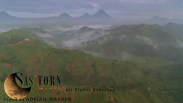 Forward tracking aerial shot, Rwandan countryside, camera passes low over farm land and mist filled valleys with exposed ridges in morning sunlight, volcanoes in background (Mt Sabyinyo, Gahinga and Muhavura)
