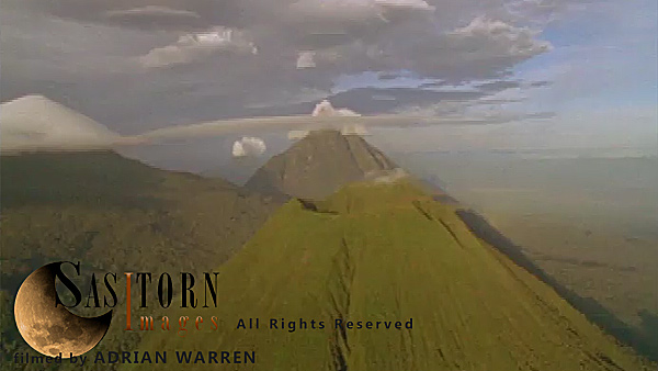 Forward tracking aerial shot, Virunga volcanoes, camera approaches Mt Visoke (Bisoke) with Mt Mikeno behind and Mt Karisimbi to left of shot, camera tilts down over crater lake and slowly tilts back up to show Mt Mikeno with cloud covered summit