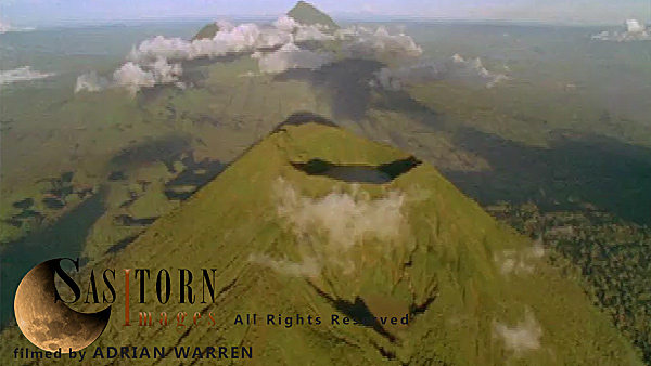 Forward tracking aerial shot, Virunga volcanoes, camera passing high over Mt Visoke (Bisoke) with Mts Sabyinyo and Gahinga in background, camera tilts down over crater lake (in shadow) camera tilts back up to show distant volcanoes surrounded by low cloud